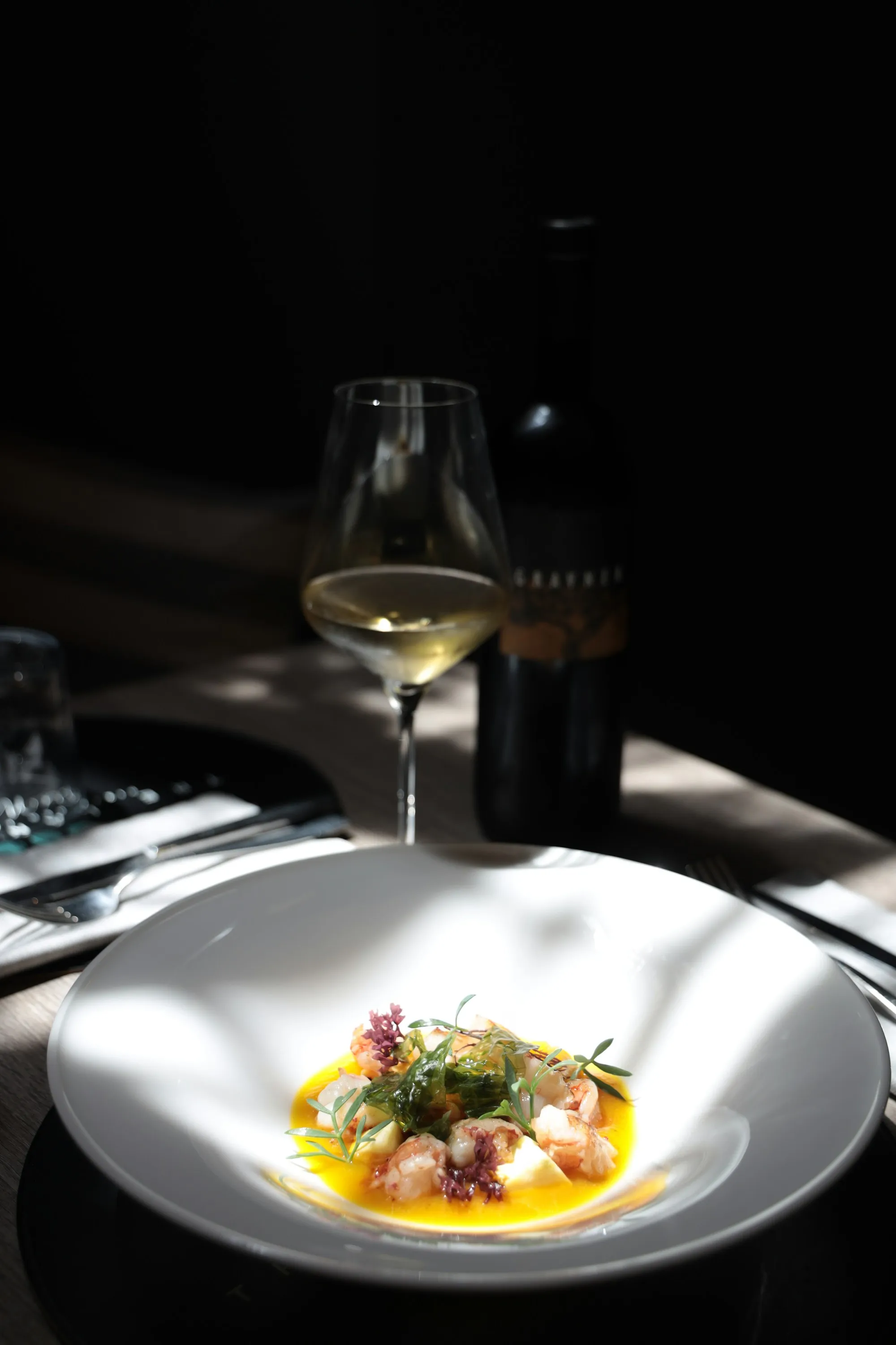 Piatto fondo in penombra con crudo di mare, crema allo zafferano ed erbe, accanto a un calice di vino bianco