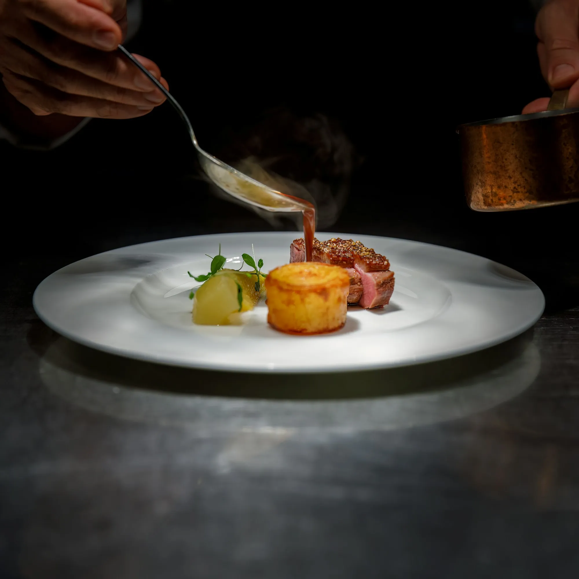 Uno chef versa una riduzione con un cucchiaio su un petto d’anatra e una patata, su fondo nero