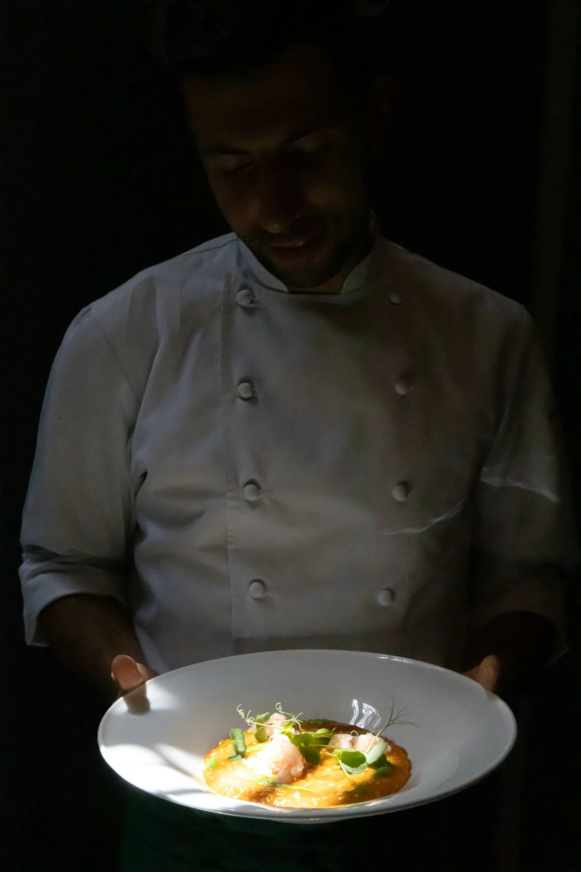 Uno chef in giacca bianca, il volto in ombra, porta un piatto di risotto illuminato nel buio