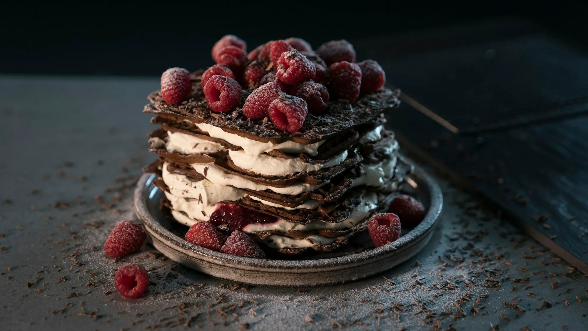 Millefoglie di cioccolato con crema e lamponi su un piatto scuro, con briciole di cacao sul tavolo