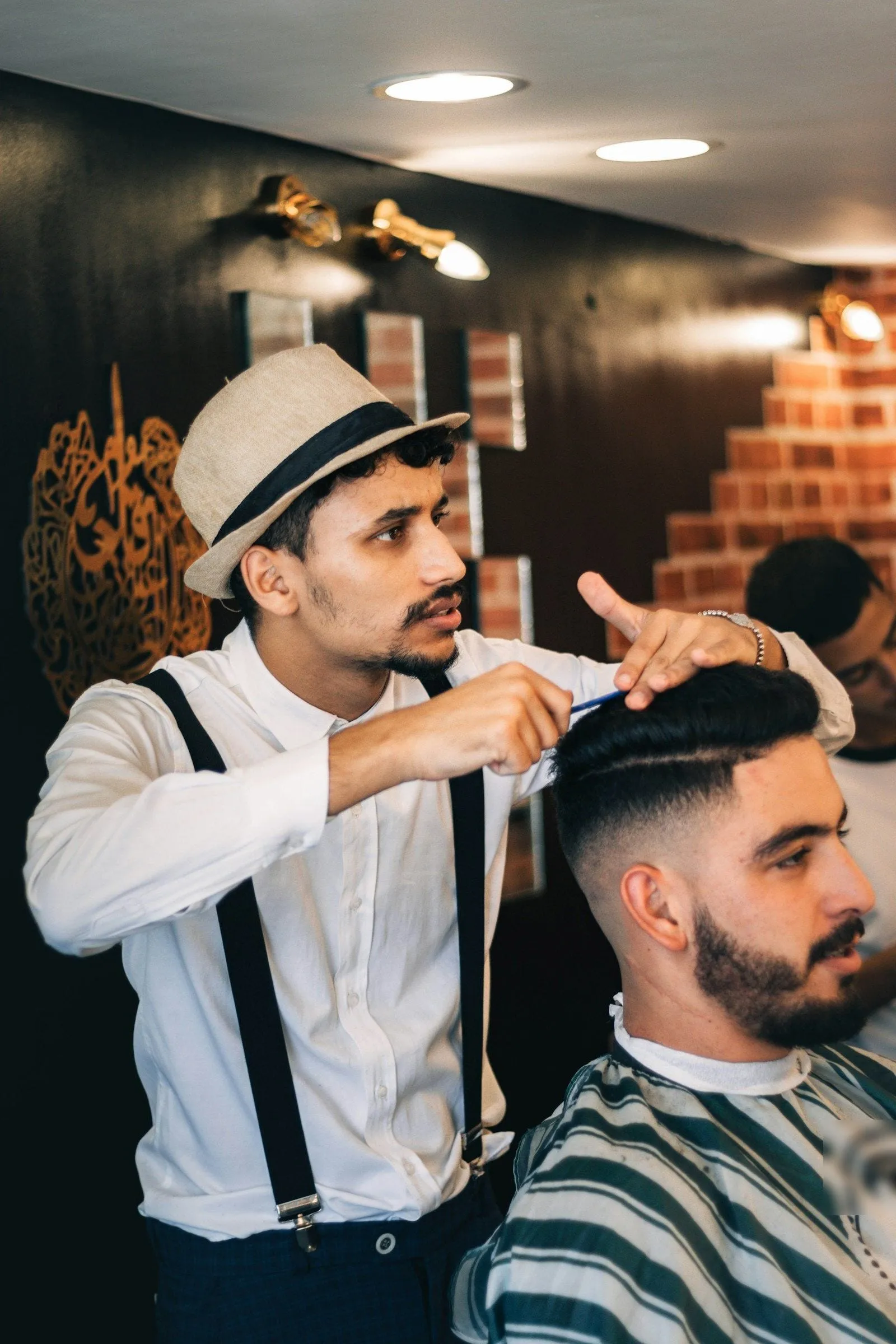 Un barbiere con cappello e bretelle sistema il ciuffo di un cliente avvolto nella mantellina a righe verdi e bianche