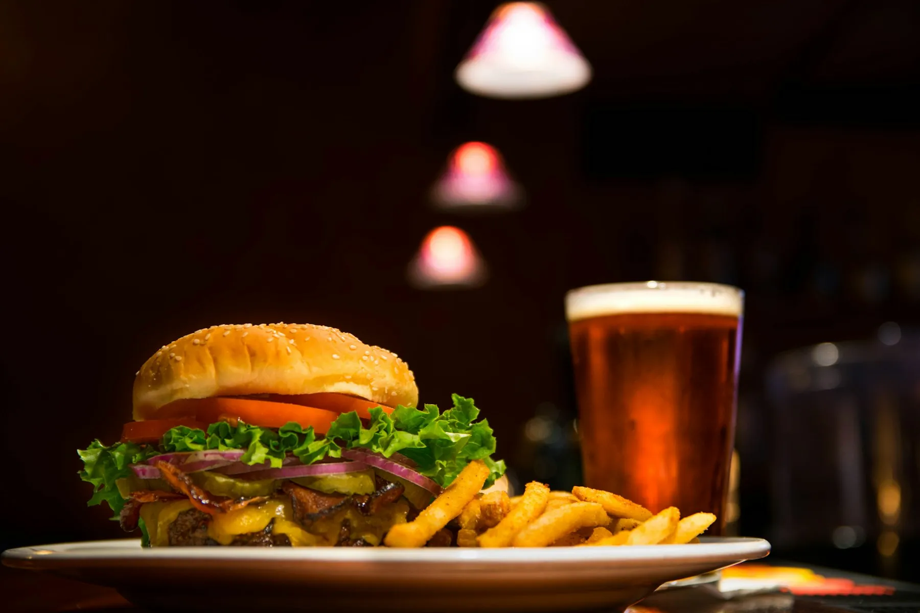 Un panino con insalata e pomodoro, patate fritte e una birra ambrata sullo sfondo.